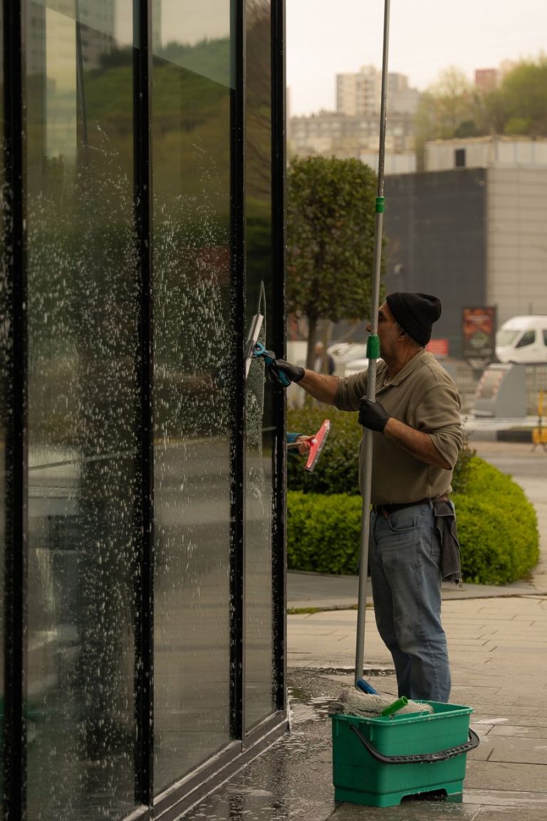 Fensterreiniger zieht eine ebenerdige Glasfront mit dem Rakel ab, daneben Teleskopstange und Eimer